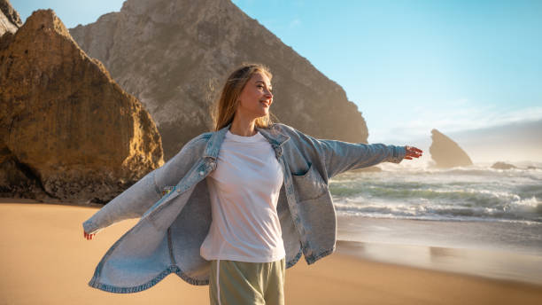 Smiling woman in denim shirt standing with arms outstretched on seashore near rocks on sunny summer day.